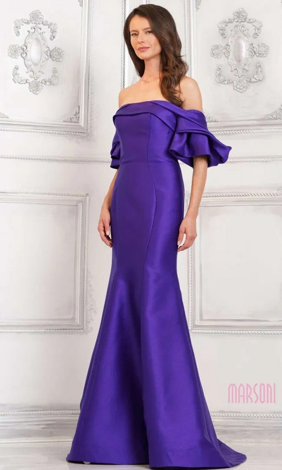 A woman poses against an ornate white wall in the Marsoni MV1290 long formal dress, featuring an amethyst color, off-the-shoulder style, mermaid silhouette with a flared skirt, and ruffled sleeves.